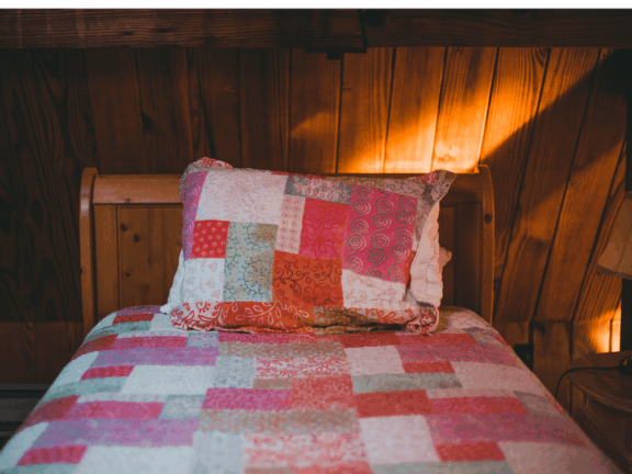 Pink patchwork quilt and pillow on a twin bed in a poorly lit wood paneled room.