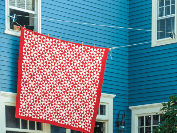 A vivid red and white patterned quilt hangs on a clothesline against a blue house.