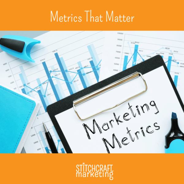 A desk covered in printouts of spreadsheets and graphs. A clipboard is on top of the papers, holding a paper that has "Marketing Metrics" written on it.